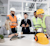 A group of construction professionals in a meeting discuss plans on a table with a tablet, blueprints, and a yellow hard hat with ear protection.