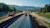 Wide view of heavy machinery paving a long stretch of road through a scenic, hilly landscape.