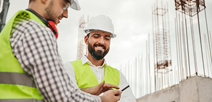 Two construction workers in safety vests and hard hats smile while looking at a smartphone on a job site with rebar structures.