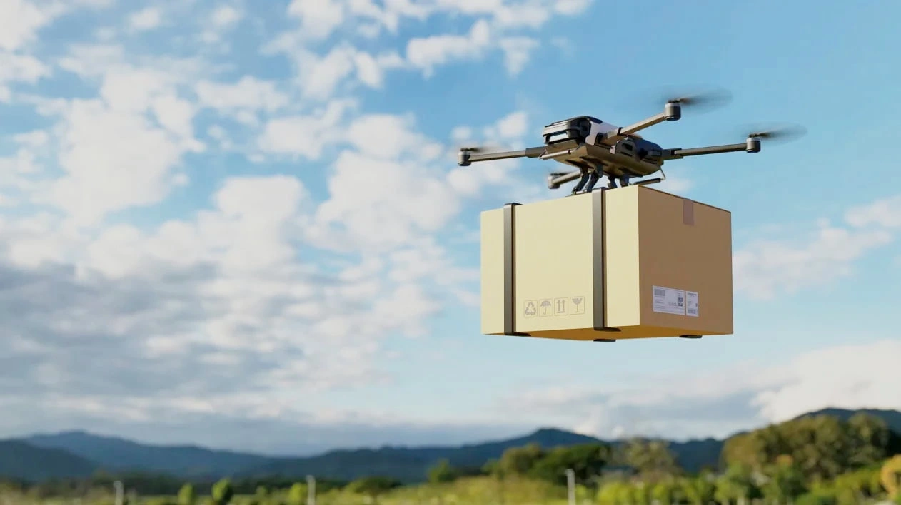 A delivery drone carrying a large cardboard box flies through a blue sky with scattered clouds over a green hilly landscape.