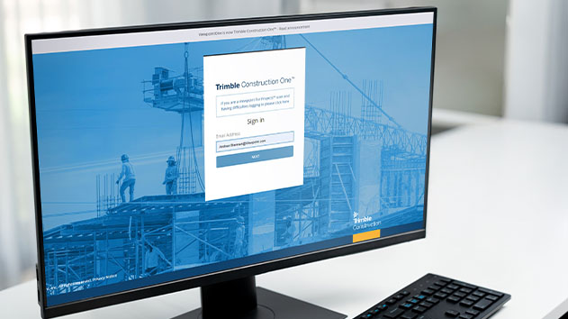  A computer monitor showing the Trimble Construction One sign-in page with a construction site background on a white desk.