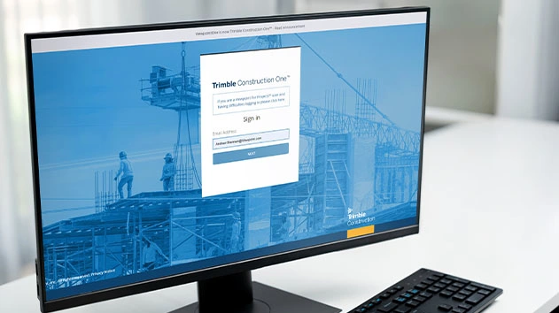 A computer monitor showing the Trimble Construction One sign-in page with a construction site background on a white desk.