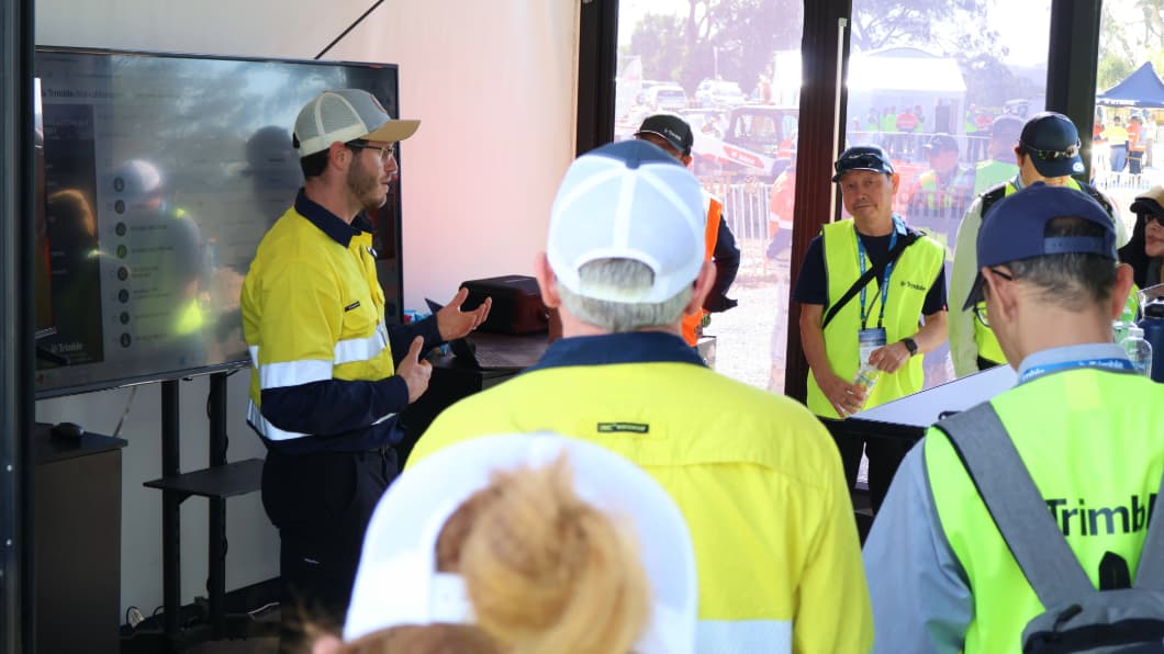 A presenter in high-visibility gear demonstrates Trimble software on a large screen for an audience at a construction industry event.