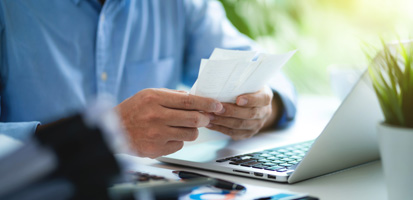 A person holds and examines papers near an open laptop on a desk. The scene conveys focus and concentration, with a blurred green background.
