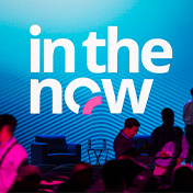 Attendees at a Trimble Dimensions conference stand before a large blue screen displaying the "in the now" event logo.