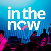Attendees at a Trimble Dimensions conference stand before a large blue screen displaying the "in the now" event logo.