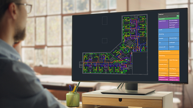  A person views a monitor displaying a complex MEP layout in Stabicad for AutoCAD, showing colorful piping and electrical systems in a building plan.