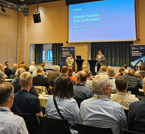 A speaker addresses a large audience at the Trimble Forestry User Conference. Attendees are seated, watching attentively and engaged.