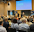 A speaker addresses a large audience at the Trimble Forestry User Conference. Attendees are seated, watching attentively and engaged.