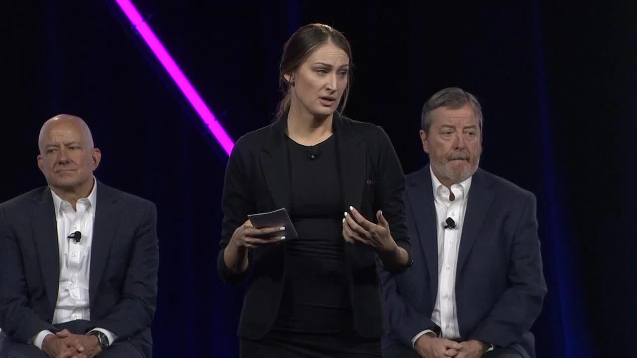 A woman speaks on stage at a Trimble event, with CEO Rob Painter and an executive seated behind her against a purple neon light background.