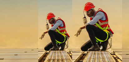 A technician in a red hard hat and safety harness kneels on a solar panel array walkway while using a handheld radio.