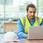 A man in a high-visibility safety vest and blue shirt uses a laptop at a table with a white hard hat in the foreground.