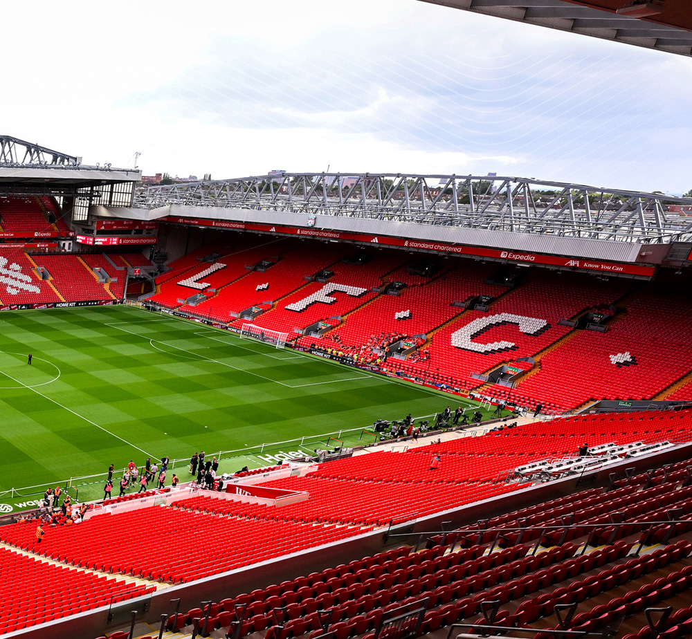 Anfield stadium empty during the day, showing vivid red stands with "LFC" spelled in white seats, a green pitch, and modern roof structures.