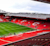 Anfield stadium empty during the day, showing vivid red stands with "LFC" spelled in white seats, a green pitch, and modern roof structures.