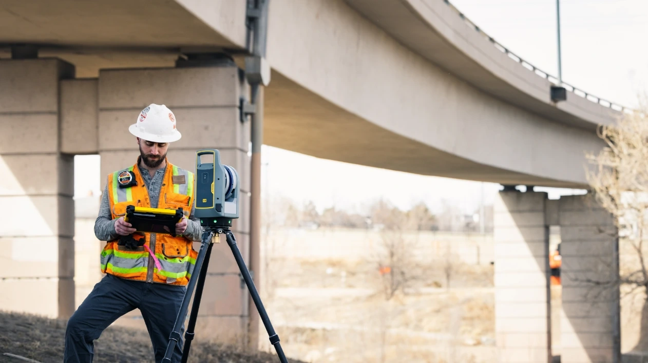 A worker in safety gear uses a Trimble X9 laser scanner and a rugged tablet to inspect a highway bridge overpass.