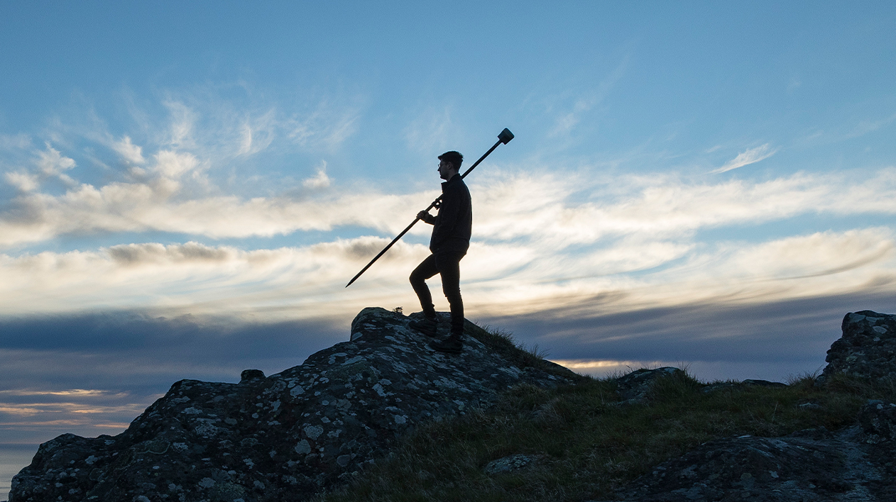 A full-body silhouette of a person standing on a rocky outcrop at dusk or dawn. The person is holding a long pole with a piece of equipment on the top, possibly surveying equipment.