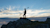 A full-body silhouette of a person standing on a rocky outcrop at dusk or dawn. The person is holding a long pole with a piece of equipment on the top, possibly surveying equipment.