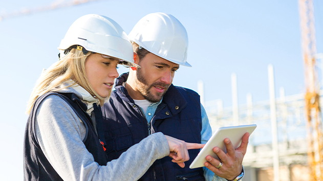 Two construction workers in safety gear review project plans on a tablet at a job site, featuring Trimble Construction One solutions.