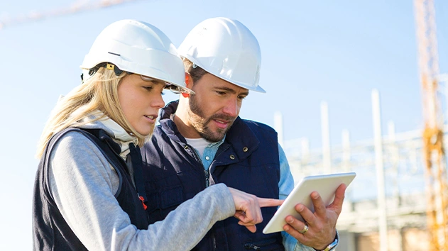 Two construction workers in safety gear review project plans on a tablet at a job site, featuring Trimble Construction One solutions.