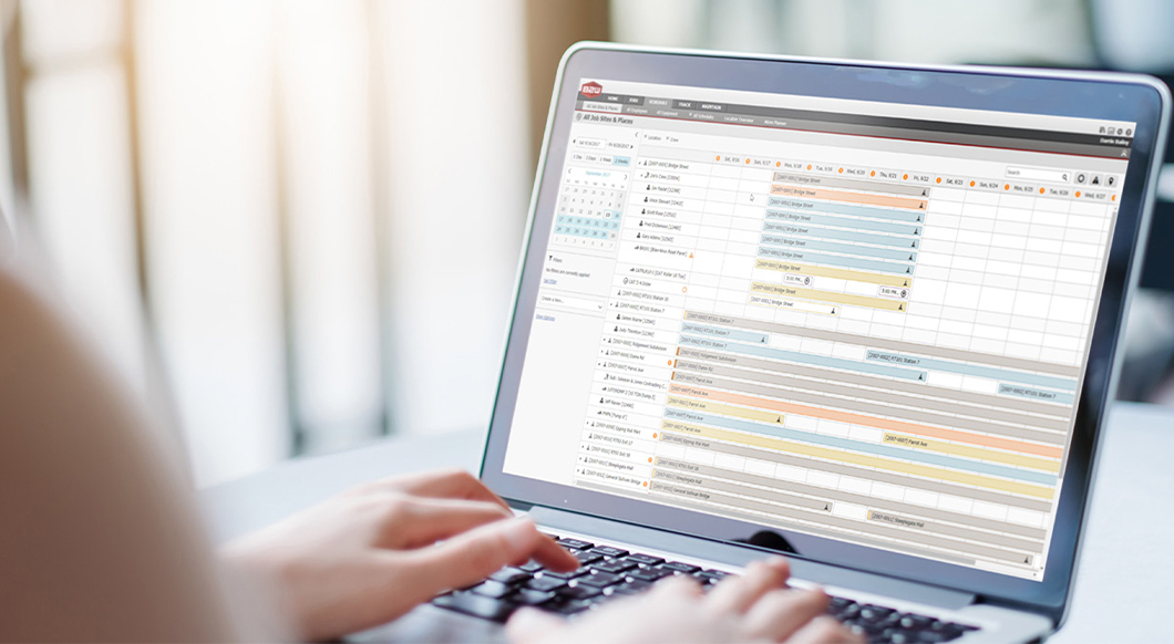 A person uses a laptop displaying the Trimble B2W Schedule interface to manage construction project resources and job site timelines.