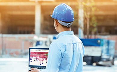 An engineer in a blue hard hat views a 3D structural model on a laptop using Trimble Tekla Structures software at a construction site.