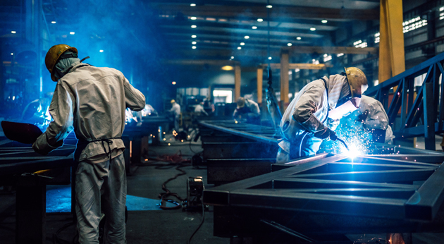 Industrial workers in protective gear weld large metal frames in a factory with bright blue lighting and flying golden sparks.