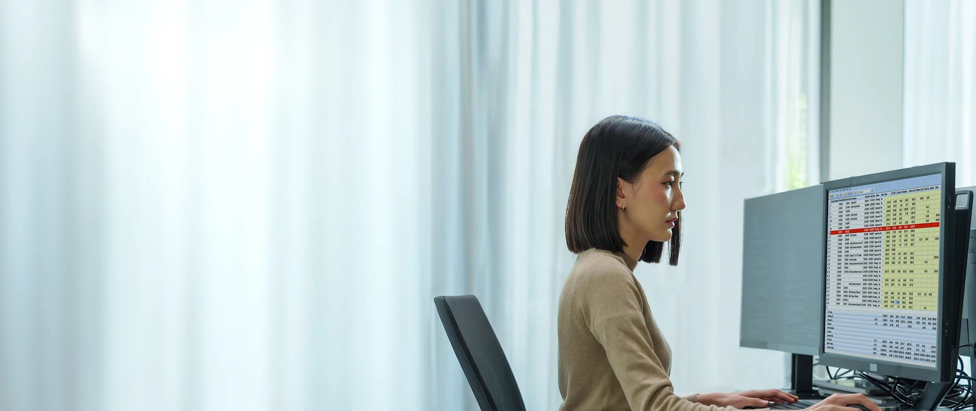 A woman sits at a desk in front of two computer monitors, looking at the screen displaying Accubid ChangeOrder software.