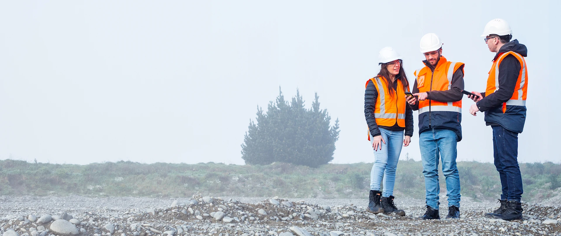 Three construction workers in safety gear use Trimble TDC600 handheld devices to collaborate on a misty, rocky job site.