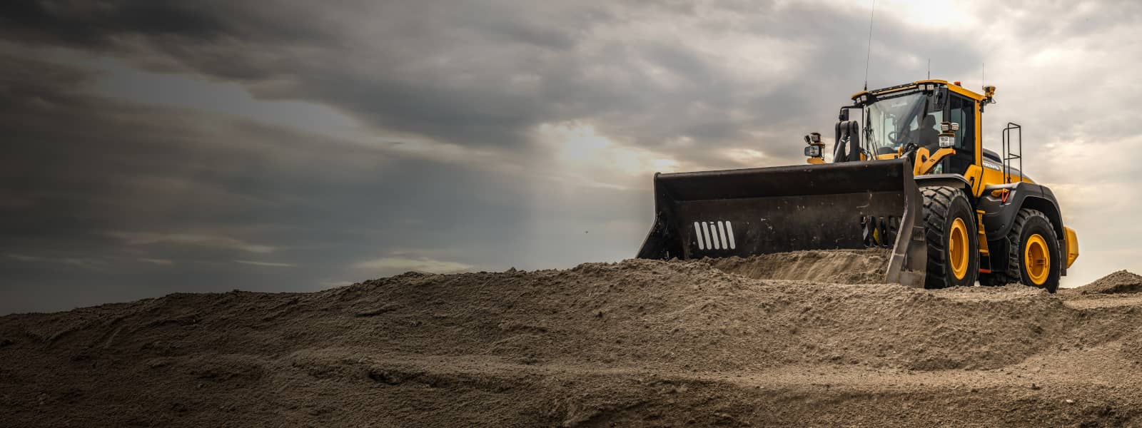 A large yellow Volvo wheel loader pushes a huge pile of sand at a construction site under a dramatic cloudy sky.