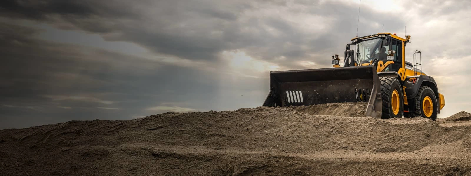 A large yellow Volvo wheel loader pushes a huge pile of sand at a construction site under a dramatic cloudy sky.