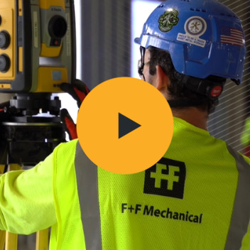 Worker in an F+F Mechanical safety vest operating a Trimble Robotic Total Station at a construction site.