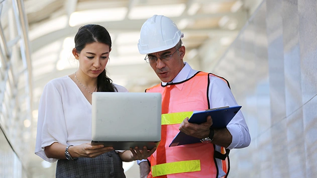 A woman and a man in a white hard hat and pink safety vest collaborate while looking at a laptop on a construction site.