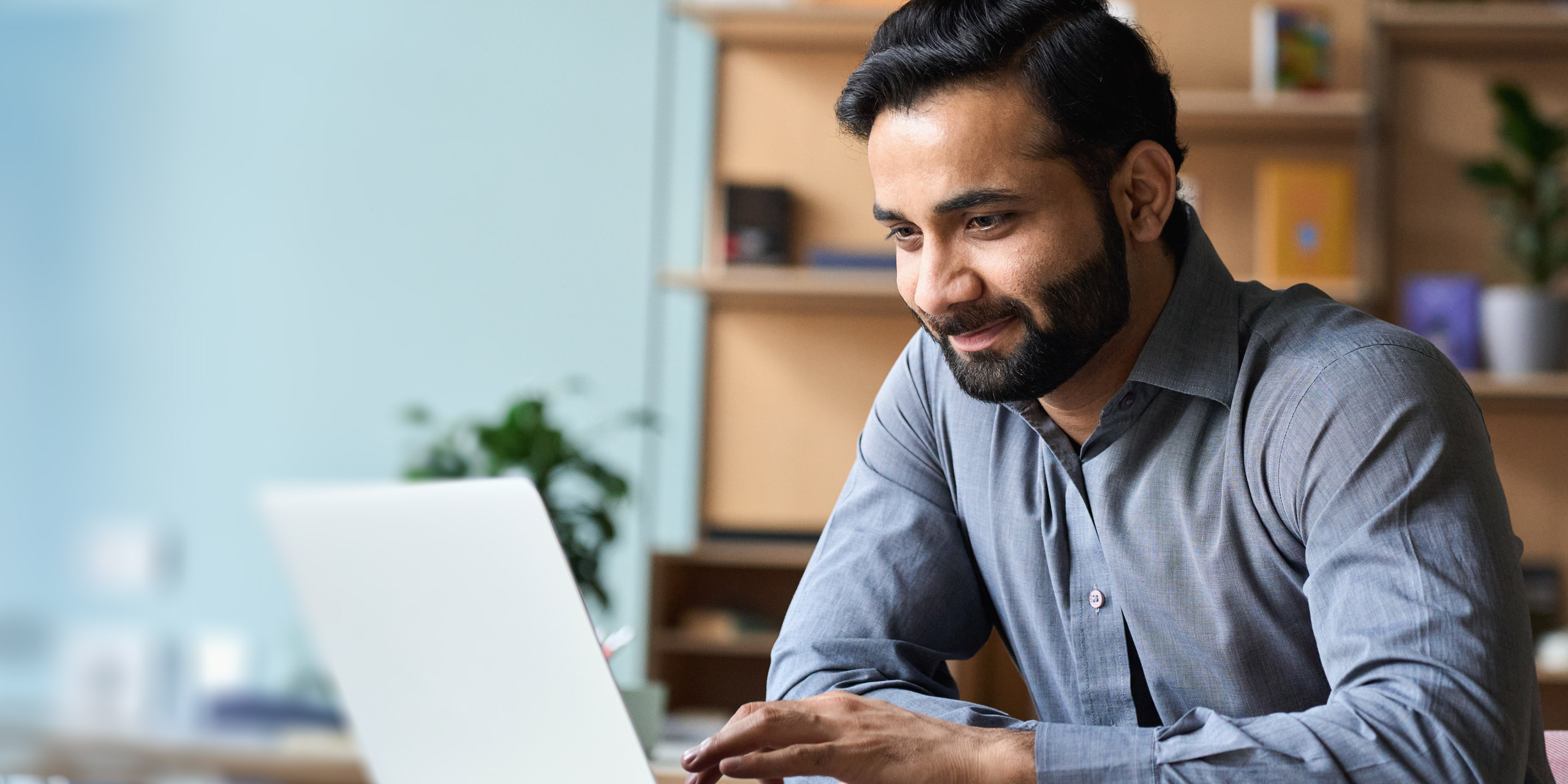 A smiling man dressed in business casual clothes looks at his laptop.