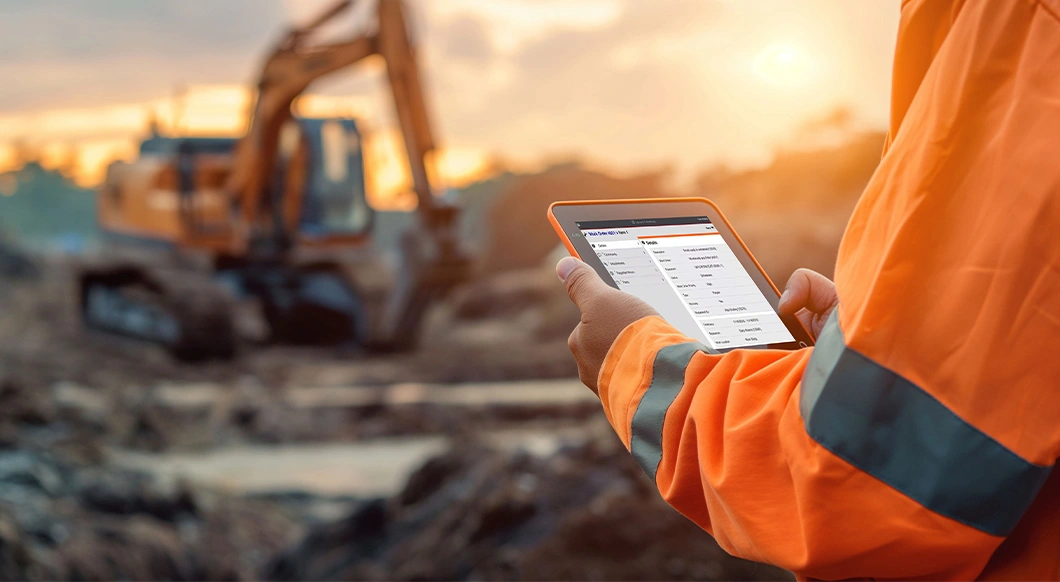 A construction worker uses a rugged Trimble field tablet showing a work order, with an excavator at a job site at sunset in the background.