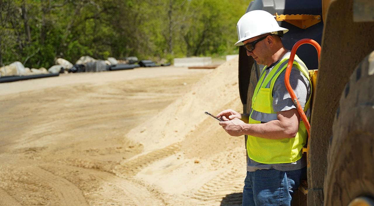 An Asian construction worker in a white hard hat and safety vest uses a tablet and stylus at a job site with concrete bridge supports.