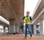 A construction engineer in a hard hat and safety vest uses a tablet to inspect the underside of a concrete bridge structure.