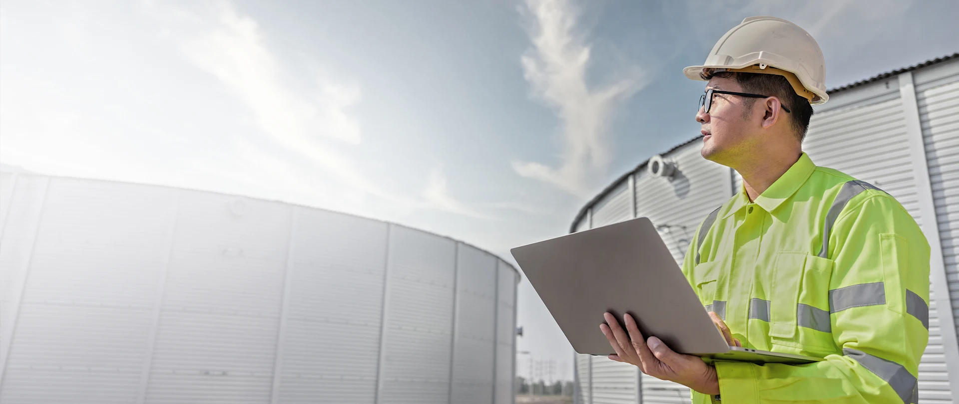 An engineer in a white hard hat and high-visibility jacket holds a laptop at an industrial site with large metal storage tanks.