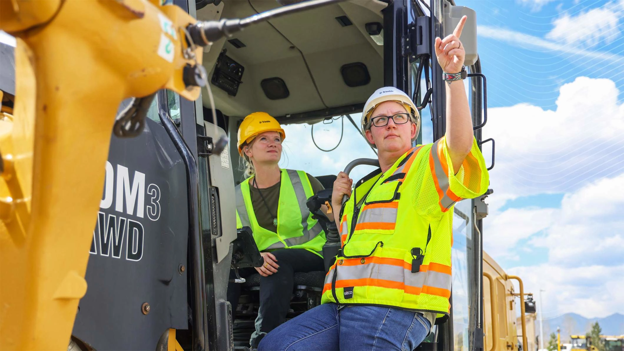 Zwei Frauen mit Trimble-Schutzhelmen stehen neben einem Cat 140M3 AWD Motorgrader. Eine zeigt zum Himmel.