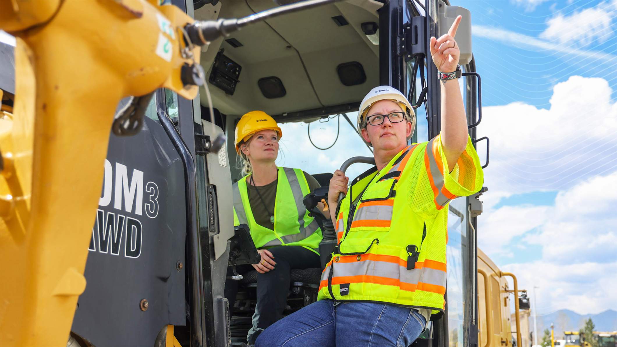 Two women in Trimble hard hats stand by a Cat 140M3 AWD motor grader; one points toward the sky.