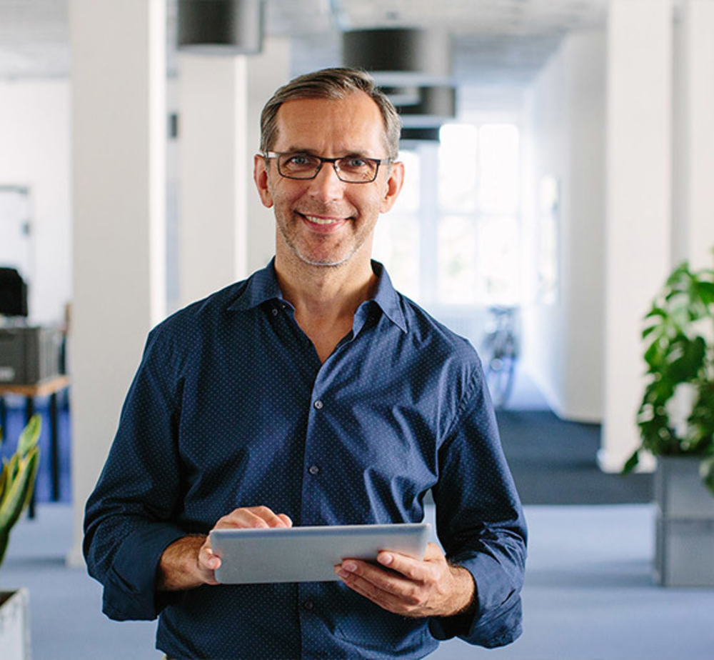 A smiling, middle-aged man wearing glasses and a blue patterned shirt holds a silver tablet in a bright, modern office setting.