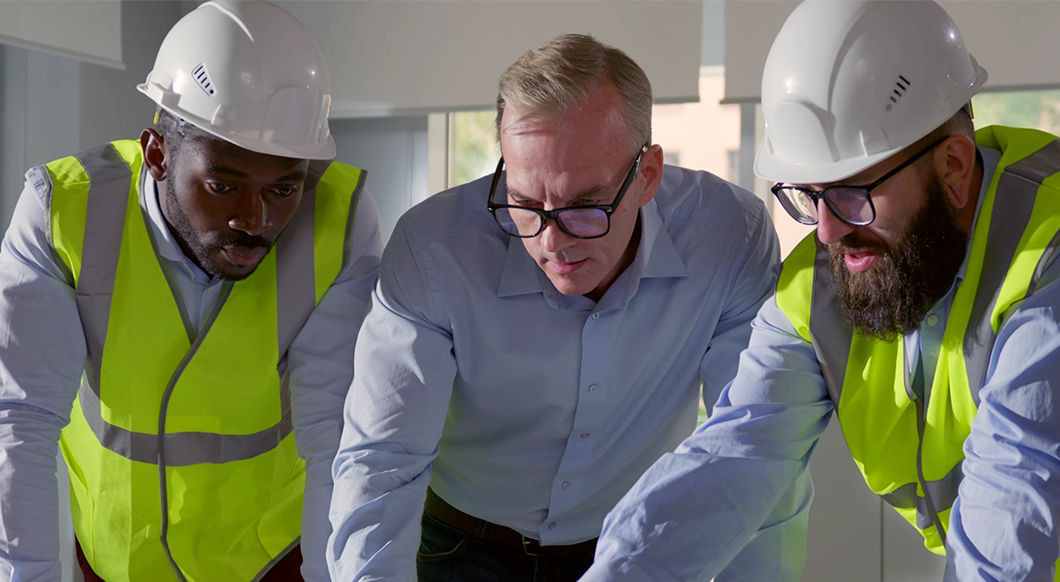 Three construction professionals, two in white hard hats and yellow safety vests, collaborate while looking down at a workspace.