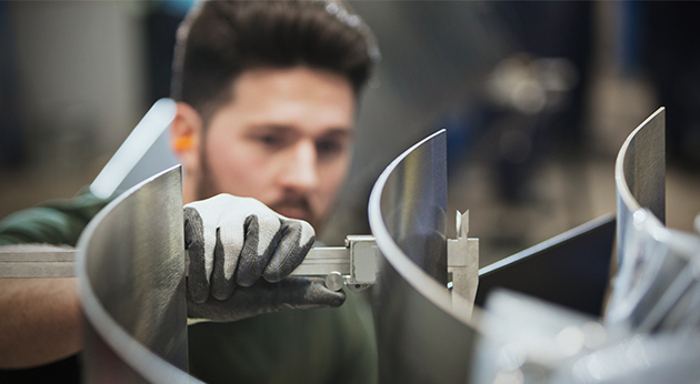 A man wearing work gloves uses a caliper to measure a large, curved metal component in a workshop.