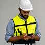 A bearded man in a white hard hat and yellow high-visibility safety vest uses a smartphone against a neutral gray background.