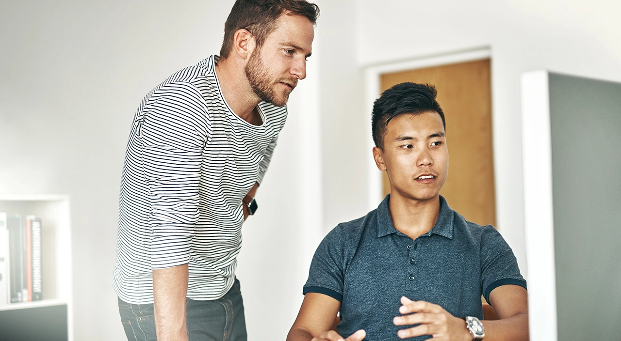 Two colleagues collaborate at a desk in an office, focused on a computer screen as they discuss their work.