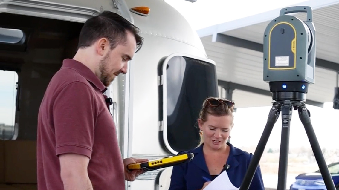 A man and woman use a Trimble T10 tablet to operate a Trimble X7 3D laser scanner on a tripod near a silver trailer.