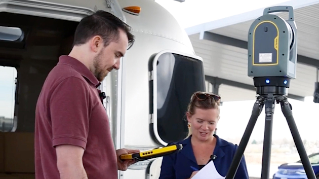A man and woman use a Trimble T10 tablet to operate a Trimble X7 3D laser scanner on a tripod near a silver trailer.