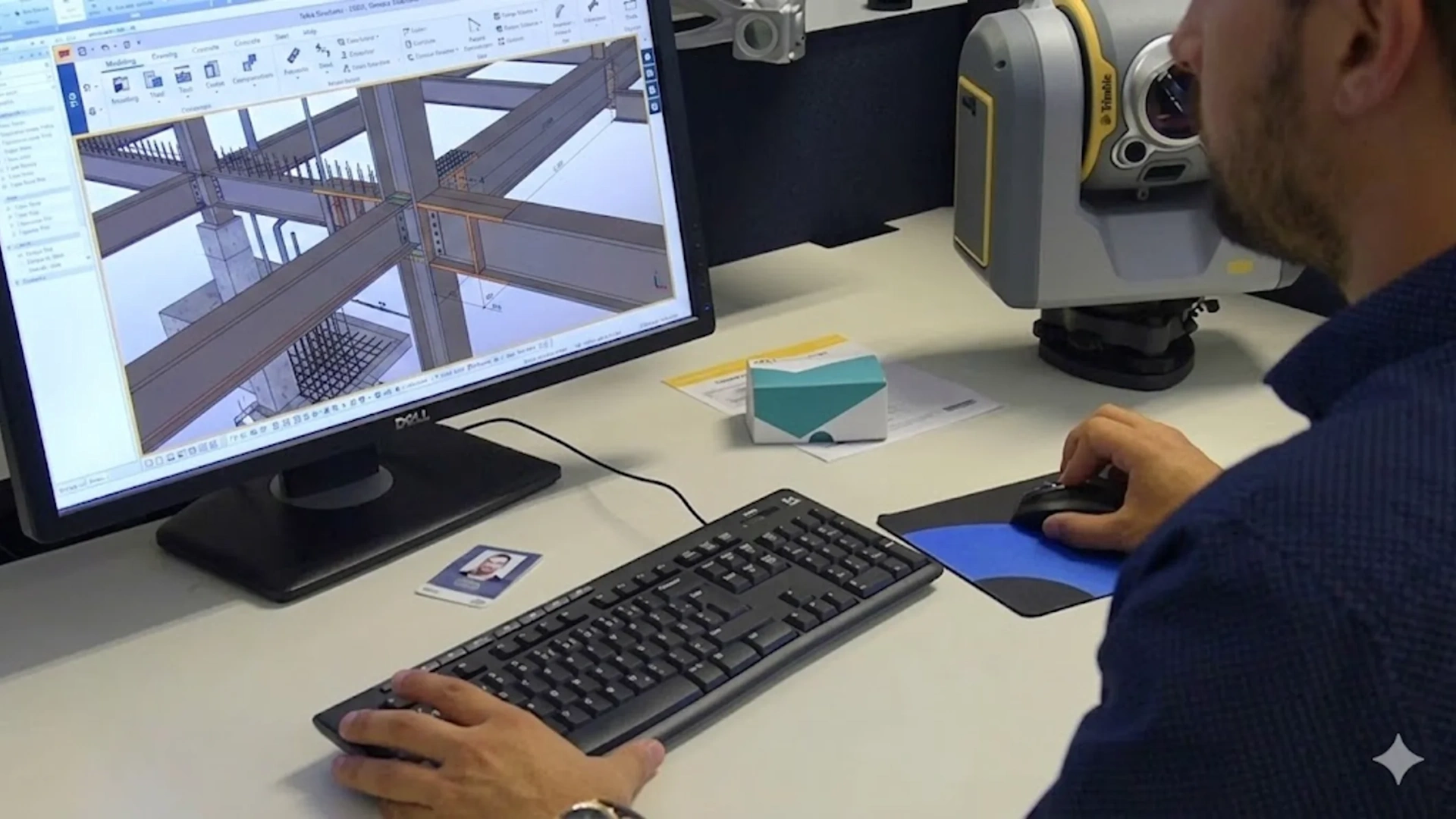 An engineer uses Tekla Structures software on a monitor next to a Trimble SX10 scanning total station on a desk.