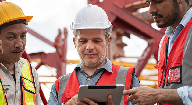 Three construction workers in high-visibility vests collaborate around a digital tablet at a job site with red steel beams.