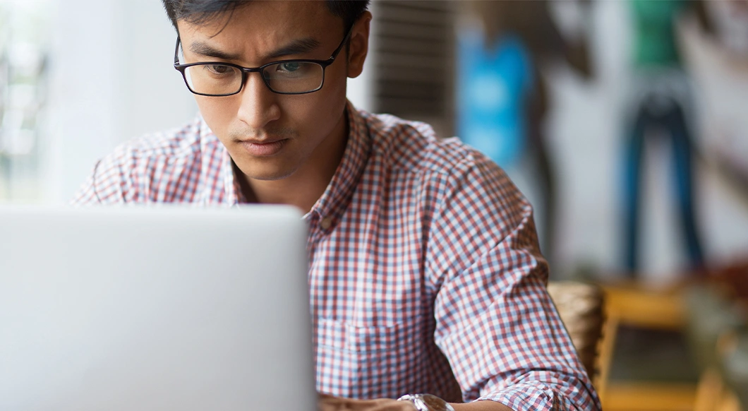 A man wearing glasses and a plaid shirt looks intently at a laptop screen in a blurred indoor setting.