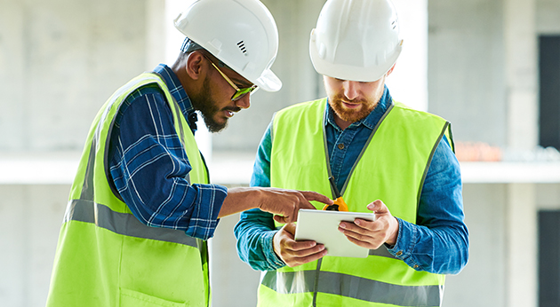 Two construction workers in hard hats and safety vests review project data on a rugged Trimble tablet at a job site.